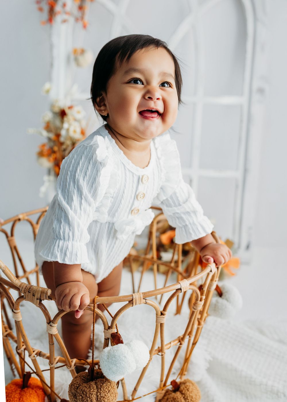 Smiling baby in white outfit sitting in a wicker basket with fall pumpkins and autumn floral decor.