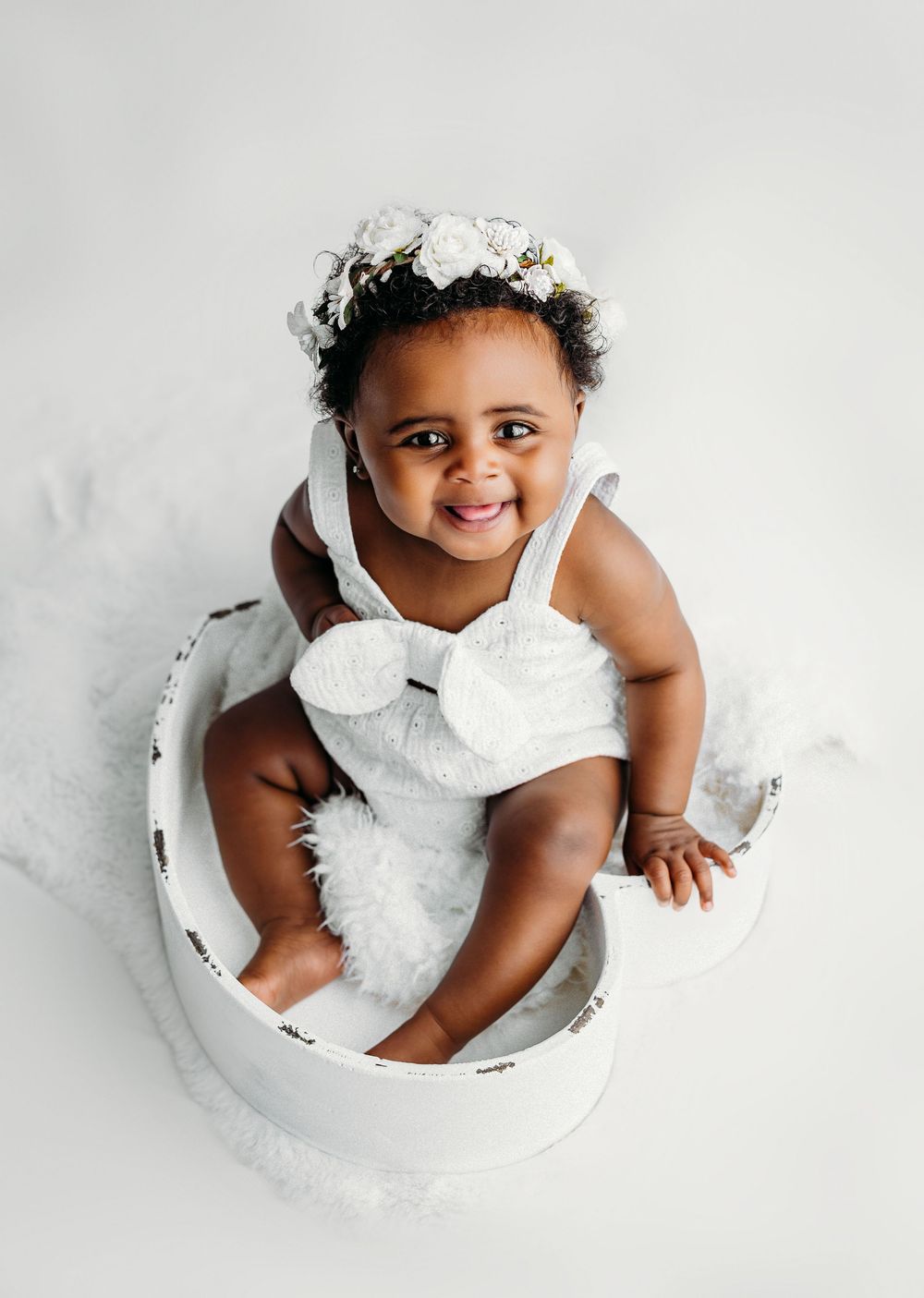 Smiling baby girl sitting in white bowl prop wearing floral crown and white outfit during professional photo session.