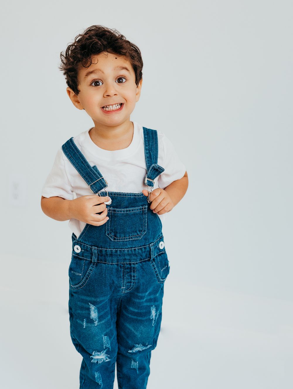 Cute toddler boy wearing denim overalls and white t-shirt, smiling and posing against a light gray background.