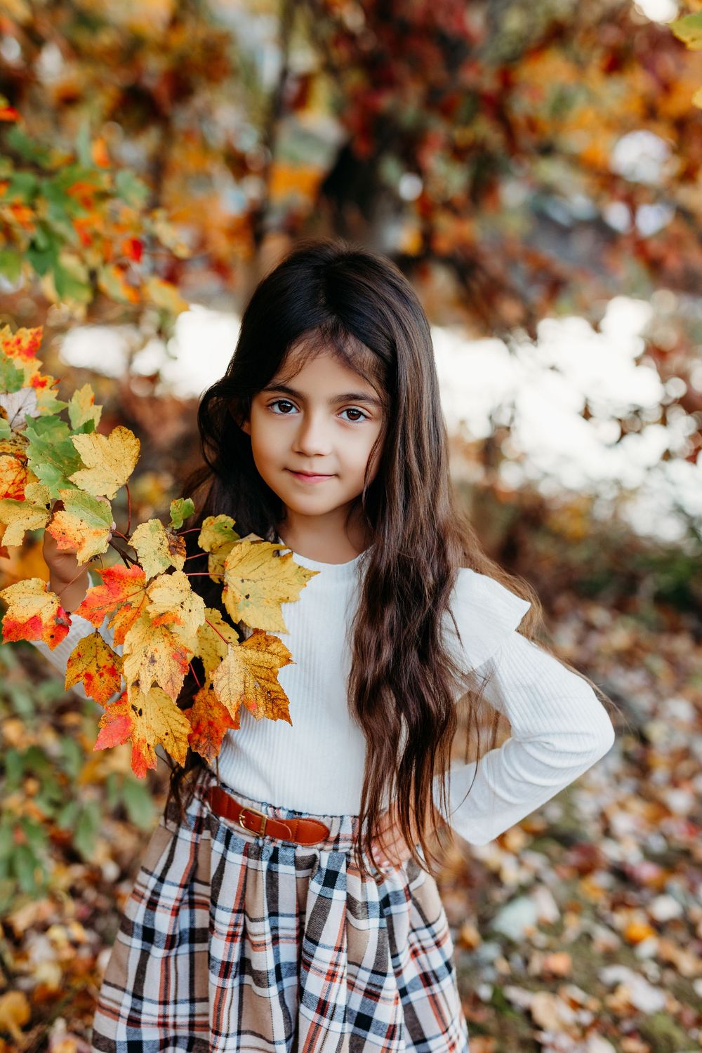 Young girl with long dark hair in white top and plaid skirt poses among colorful autumn leaves in a fall forest.