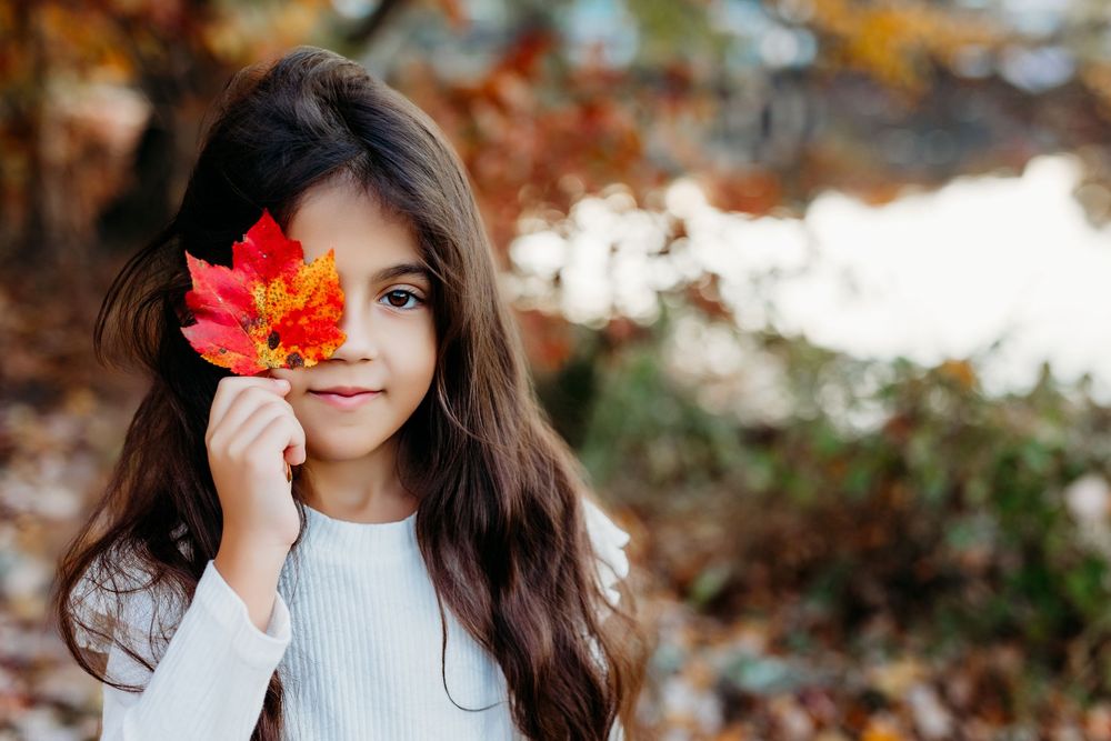 A young girl with long dark hair holds a vibrant red maple leaf beside her face in an autumn outdoor setting.
