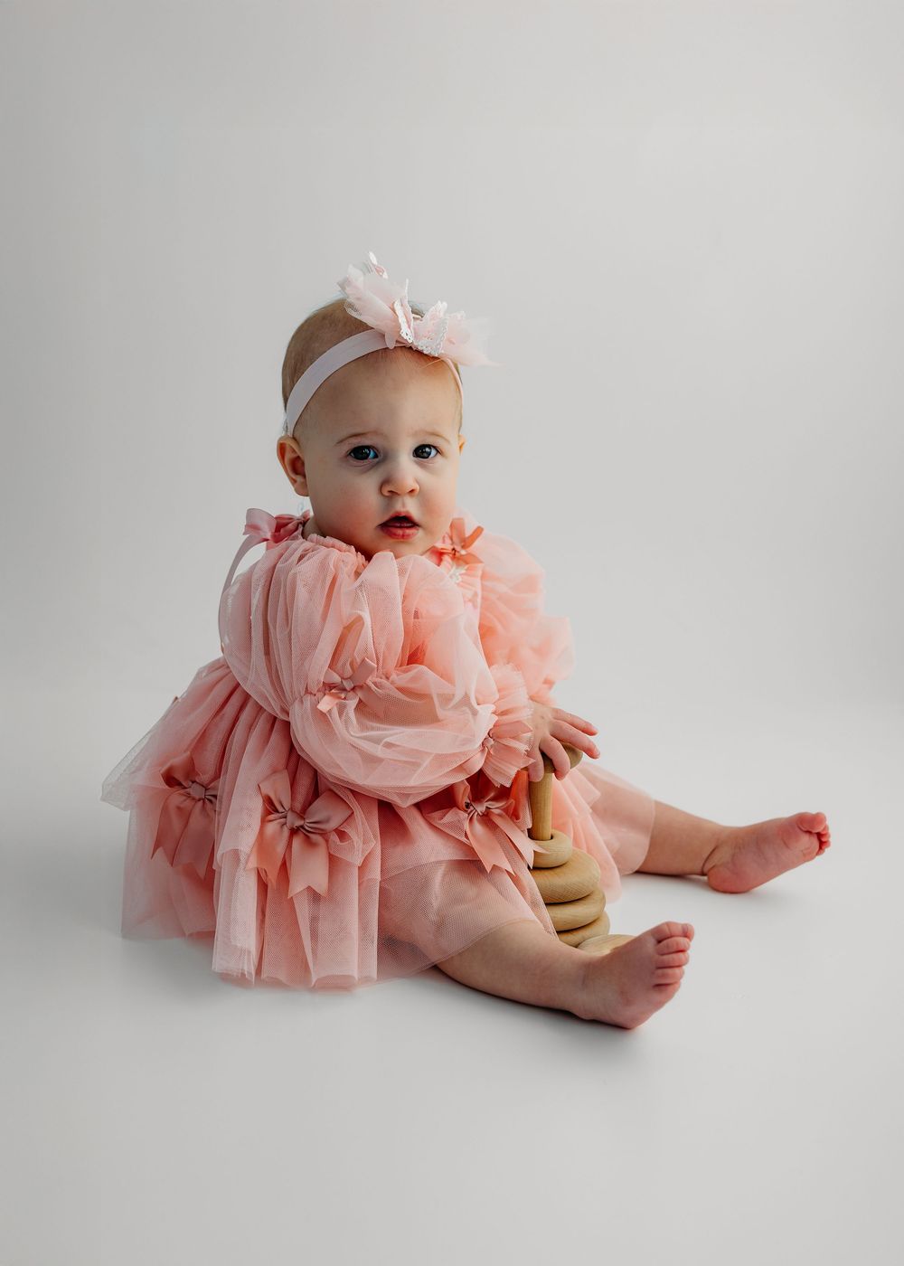 Baby girl in pink tulle dress and headband sits barefoot holding a wooden toy against white background.