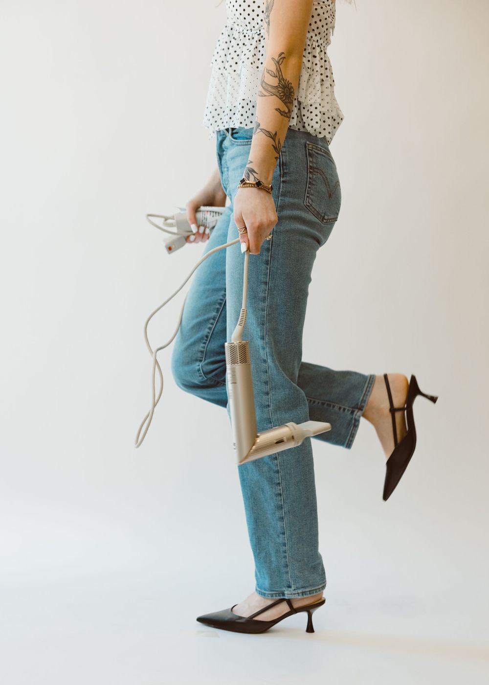 Stylish woman in jeans and heels holds a hair straightener while posing against white background.