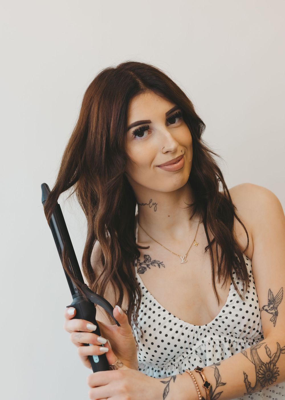 Tattooed female hairstylist holding a curling iron with long brown wavy hair wearing a polka dot top.