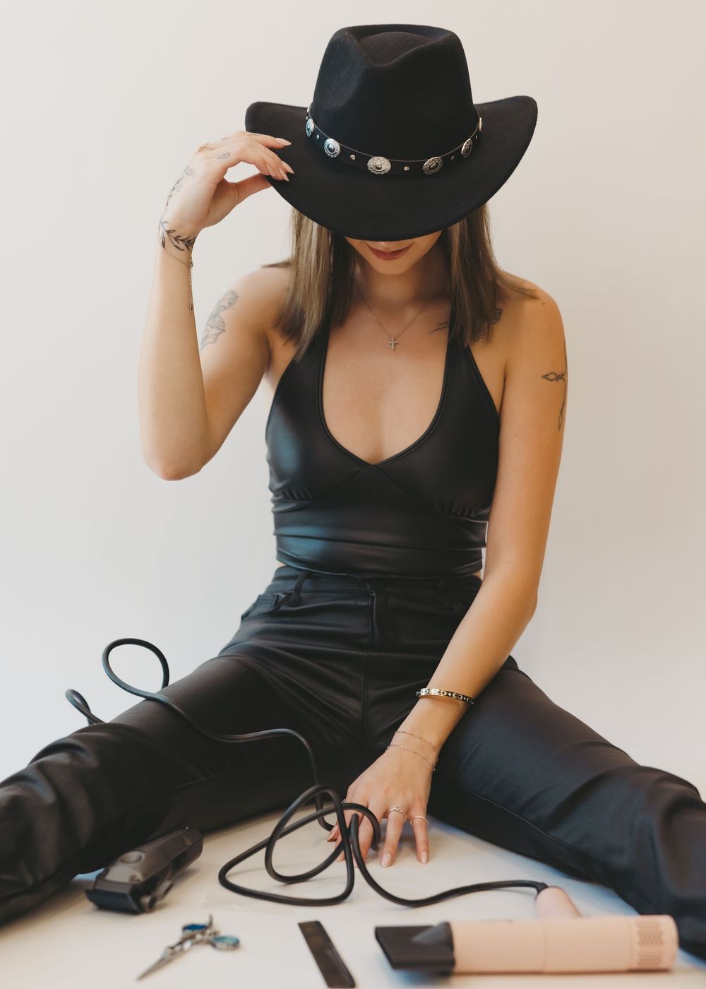 Woman in black bodysuit and leather pants wearing cowboy hat sitting with hair tools.