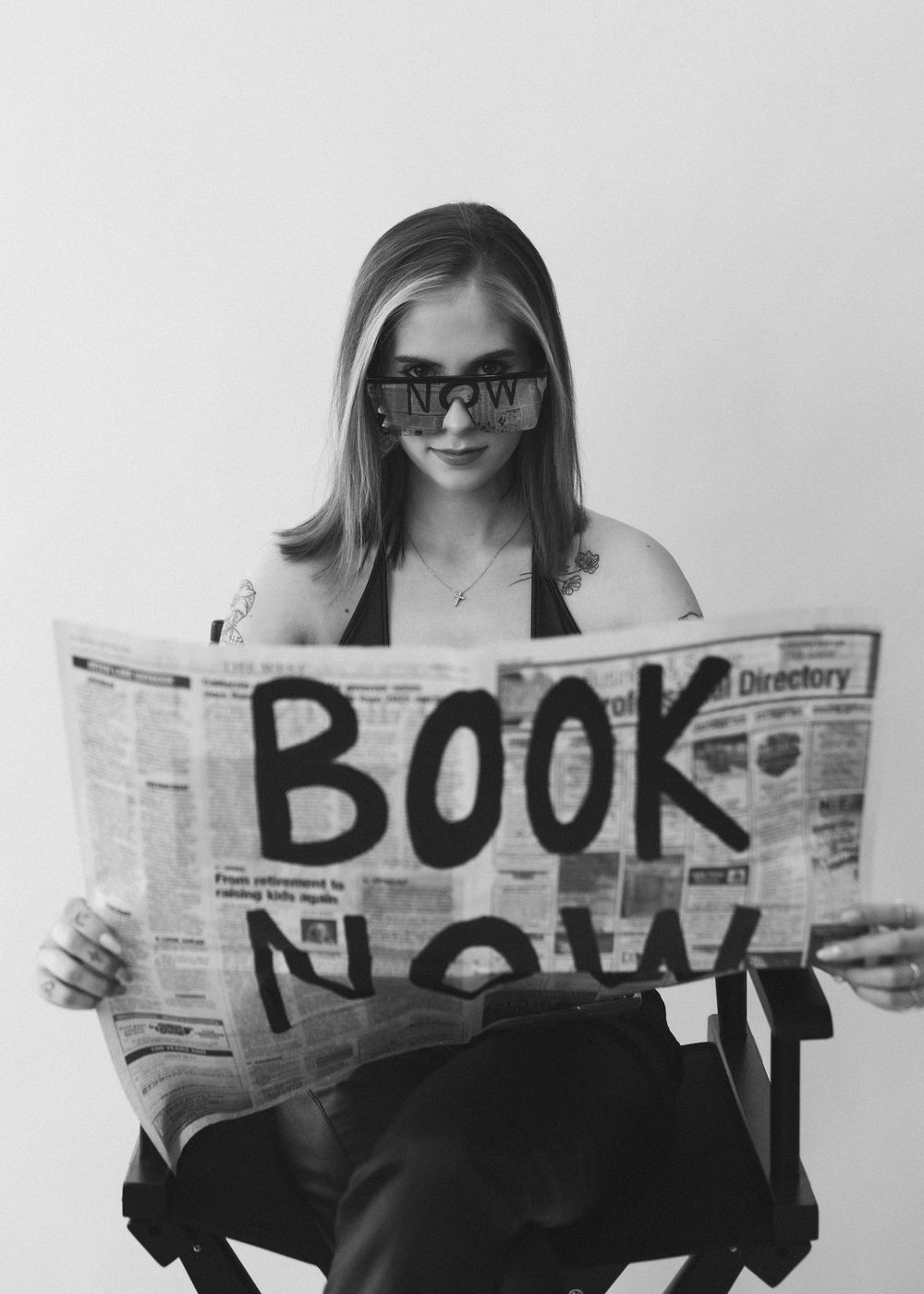 Blonde woman reading newspaper with Book Now written on it in black and white photo.