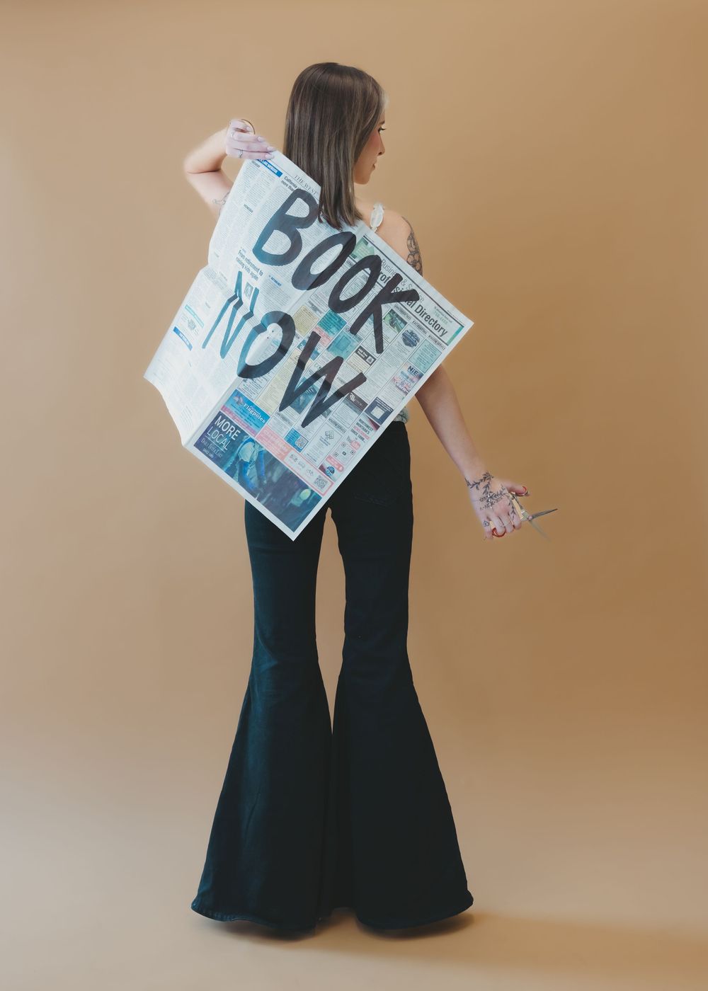 Woman holding large Book Now newspaper sign wearing wide-leg pants against beige background.