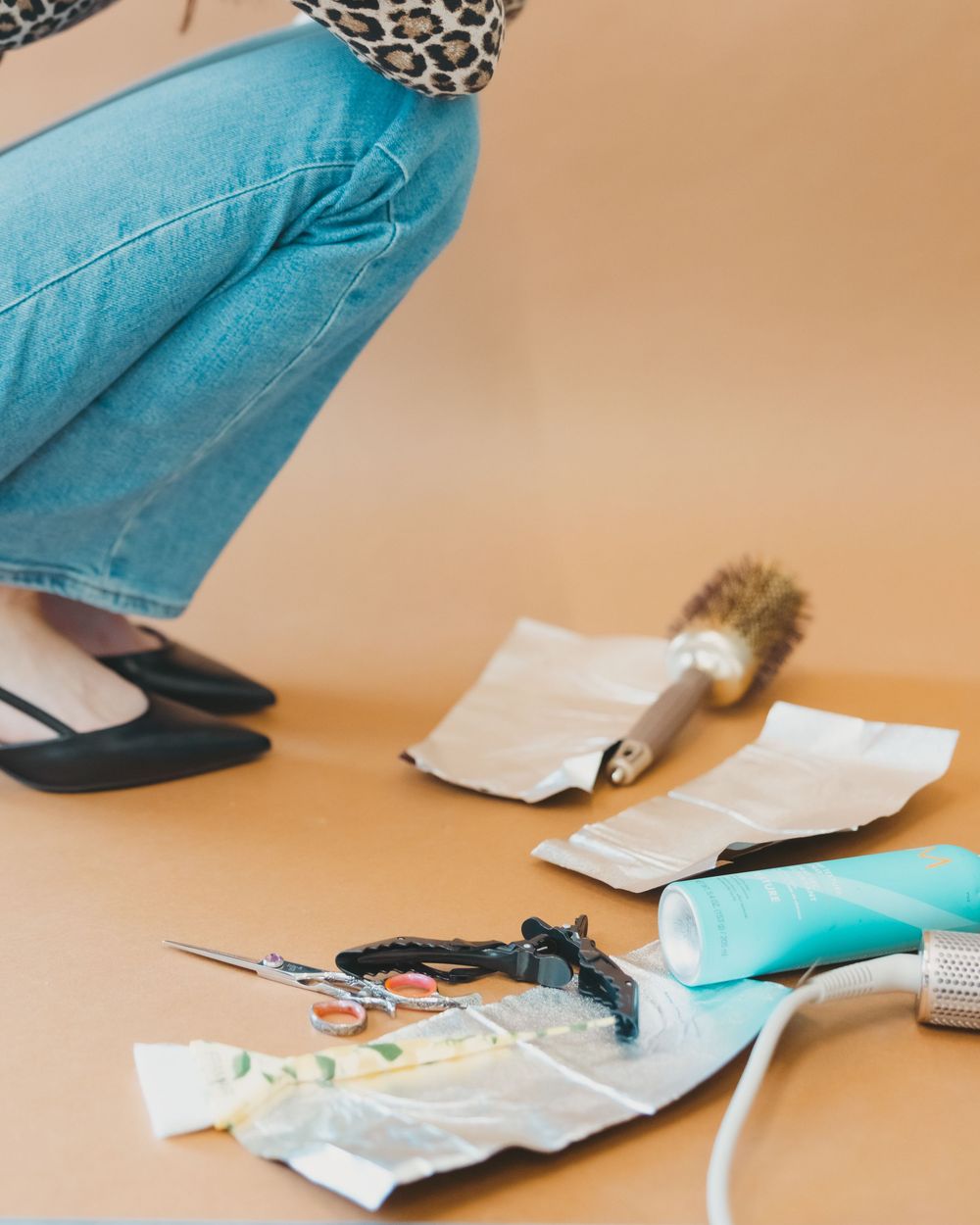 Person crouching near hair styling tools including scissors and brush on orange background.