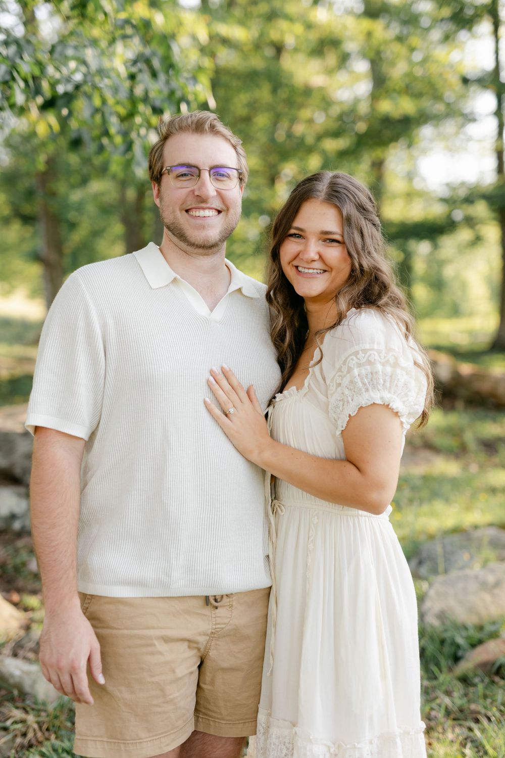 Smiling engaged couple posing outdoors in matching white and khaki outfits among green trees.