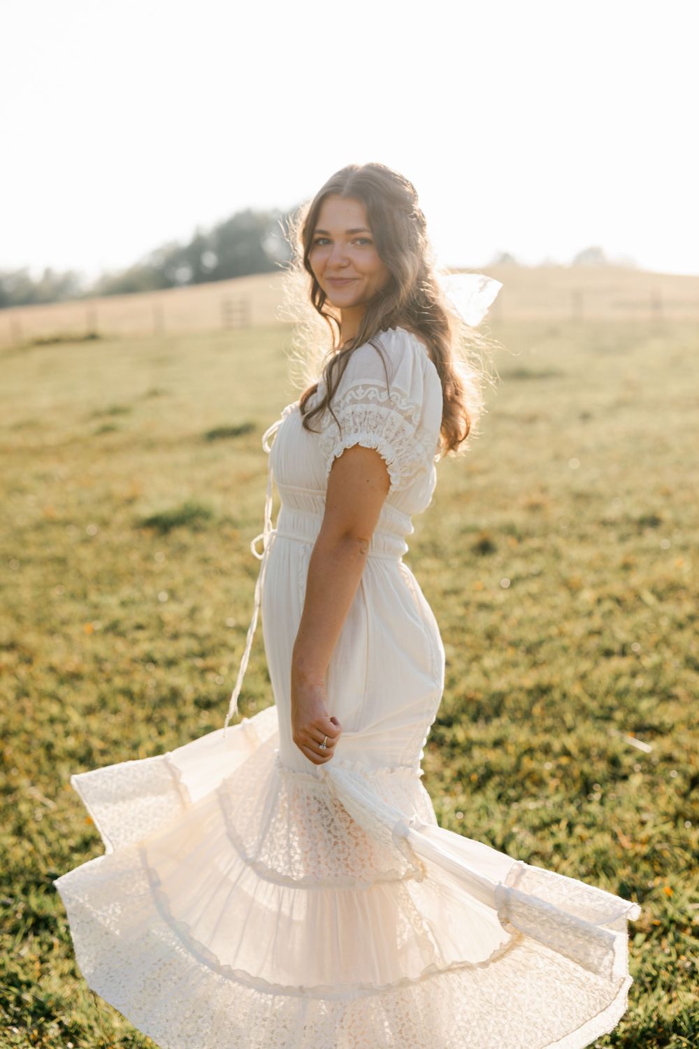 Woman in white lace midi dress twirling in a sunlit green field at golden hour.
