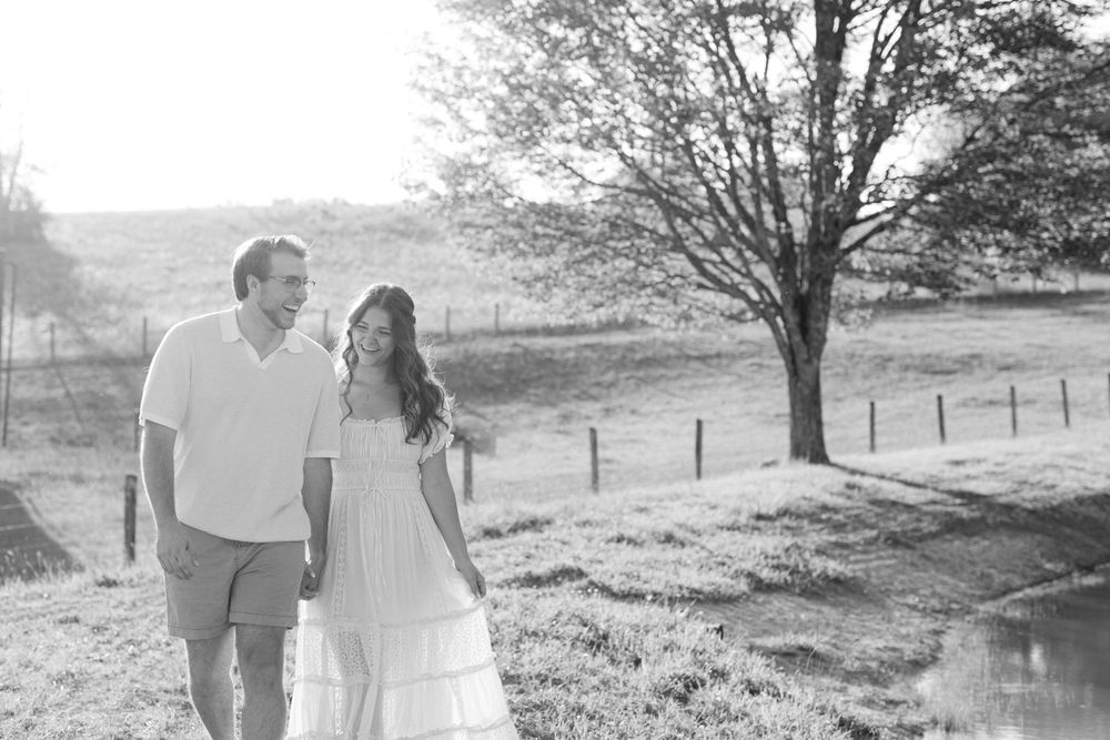 Black and white photo of a couple walking hand in hand through a scenic countryside field.