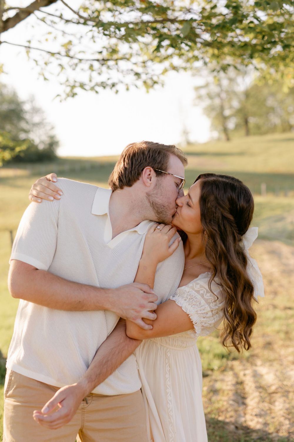 Couple sharing a romantic kiss under a tree in warm golden sunlight in an open green field.