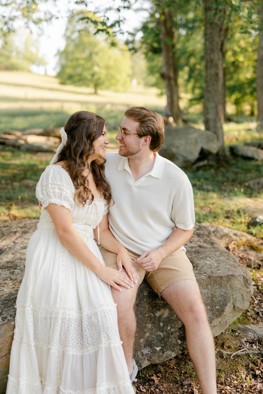 Couple sharing a tender moment on rocks in a sunlit forest, woman in white dress and man in cream polo.