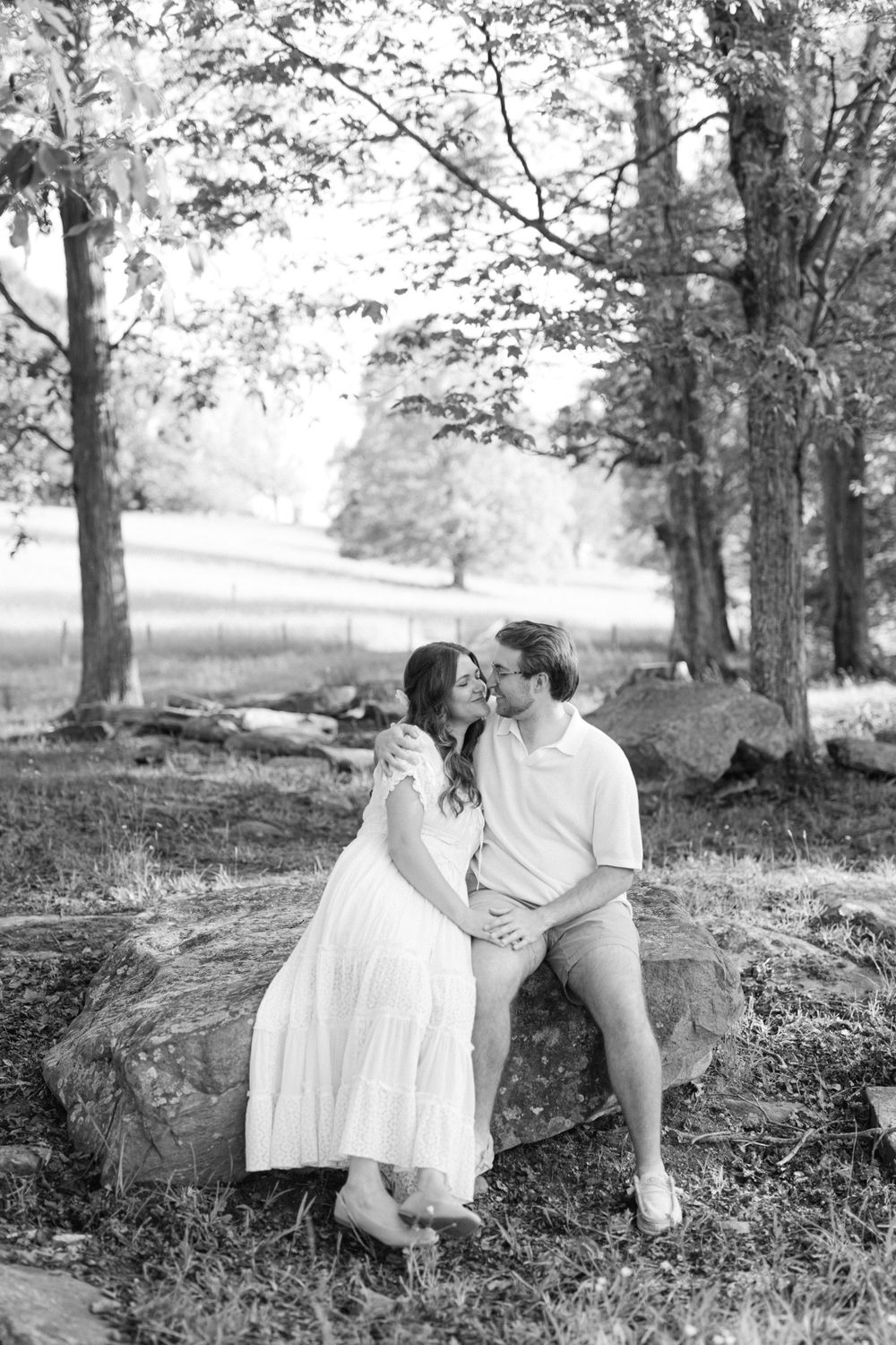Black and white photo of a couple sitting on rocks in a wooded park, sharing a romantic close moment.