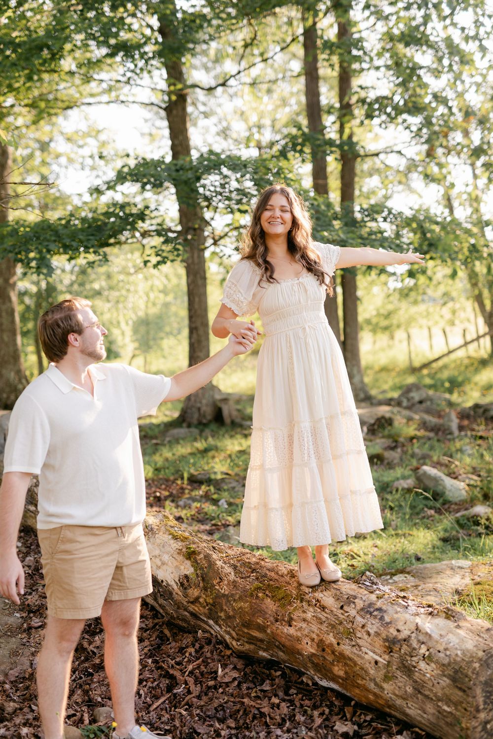 Joyful woman in white dress balancing on a log while her partner holds her hand in a sunny forest.