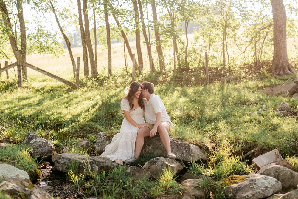 Romantic couple sharing a kiss while seated on rocks beside a small stream in a sunlit woodland.