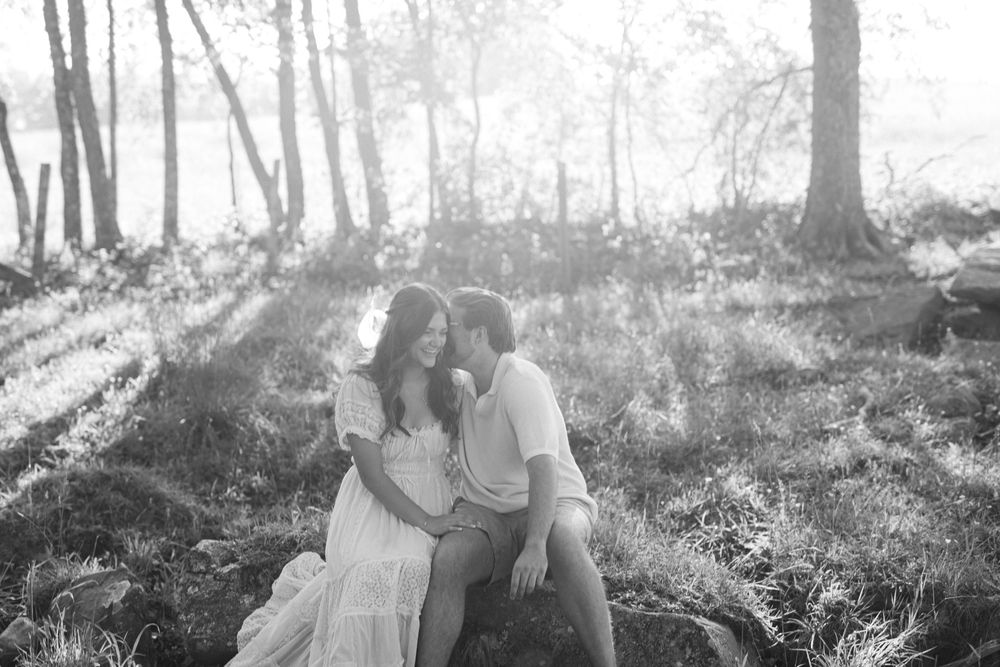 Black and white image of a couple sitting close together on rocks in a glowing sunlit forest setting.