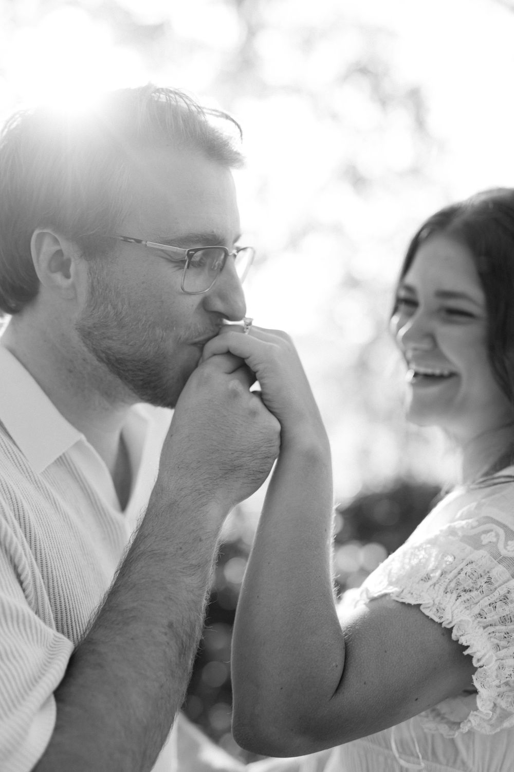 Black and white close-up of a man kissing a womans hand as she laughs joyfully in bright sunlight.