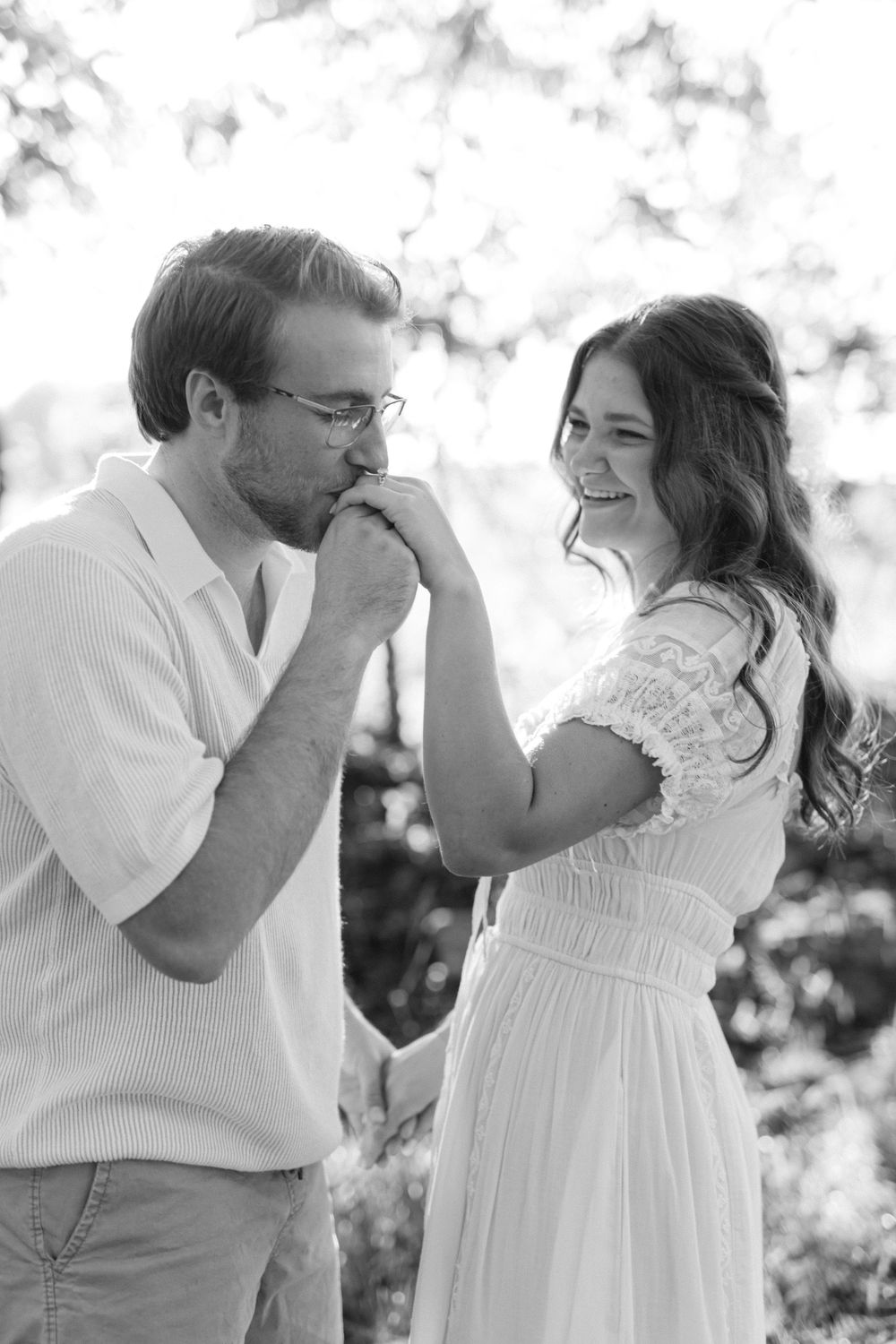 Black and white photo of a man kissing a smiling womans hand outdoors in soft natural backlit light.