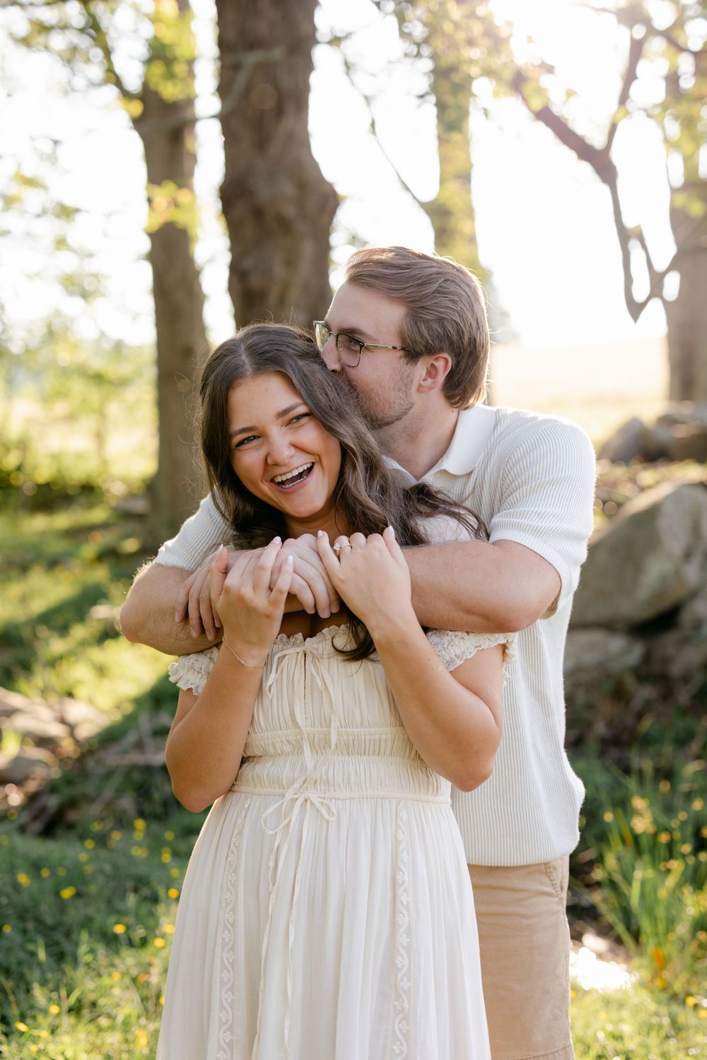 Man hugging woman from behind and kissing her cheek as she laughs joyfully in a sunny forest setting.