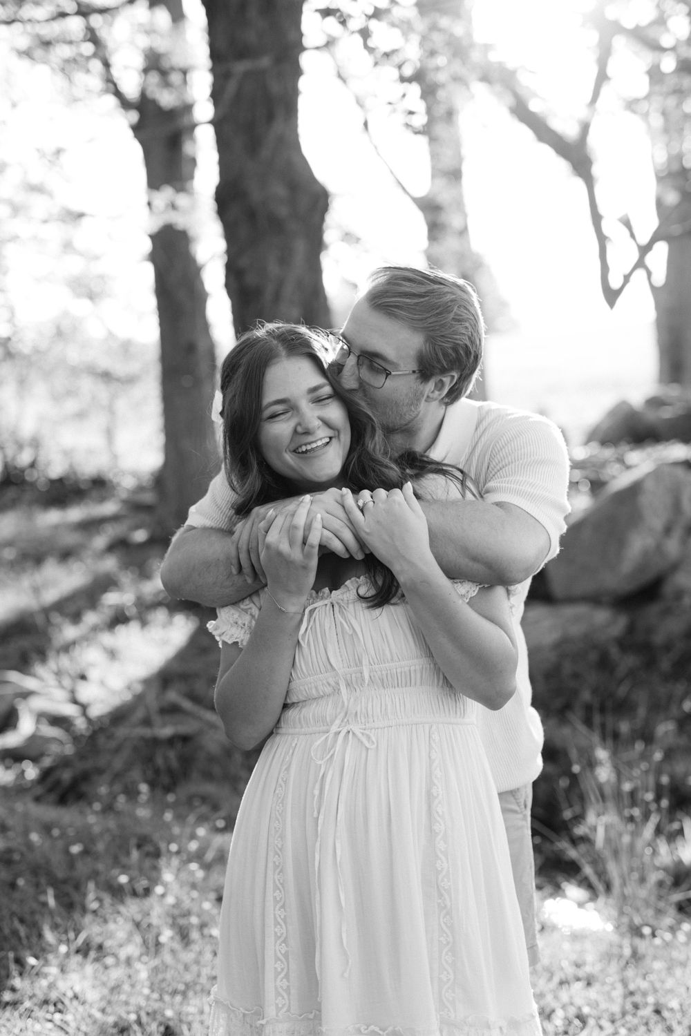 Black and white photo of a couple embracing outdoors, man kissing woman's cheek as she smiles joyfully.