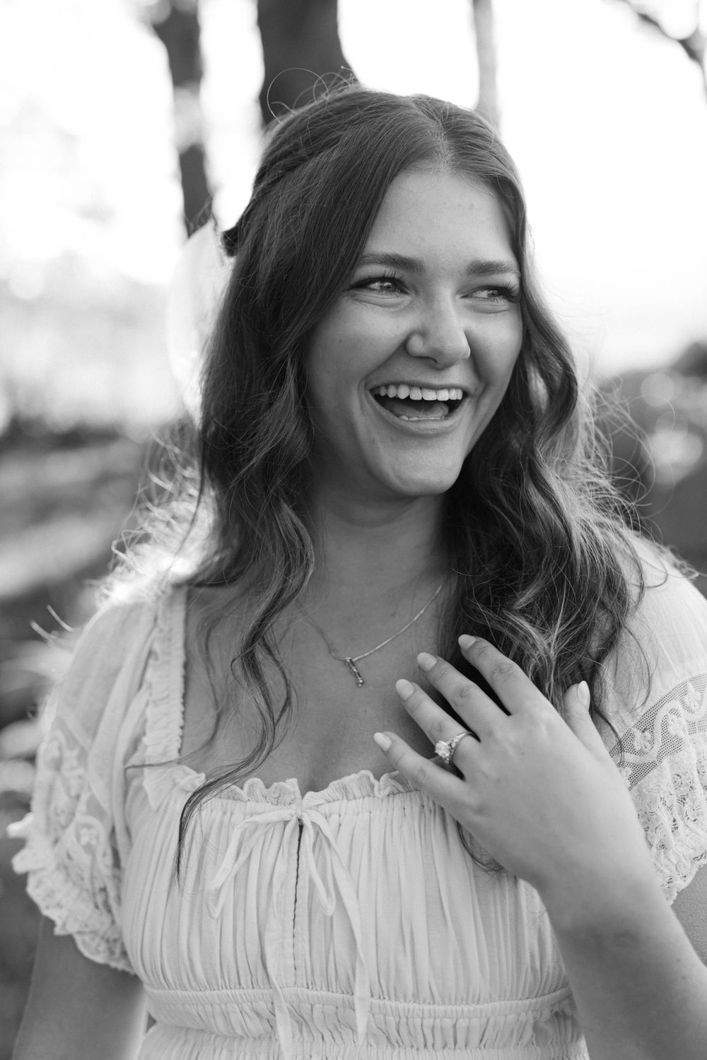 Black and white portrait of a laughing brunette woman in white dress displaying her engagement ring.