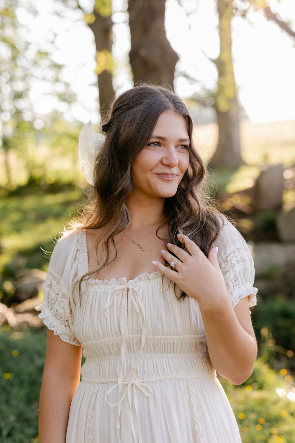 Young brunette woman in cream lace dress smiling softly outdoors in golden sunlight with hair bow.