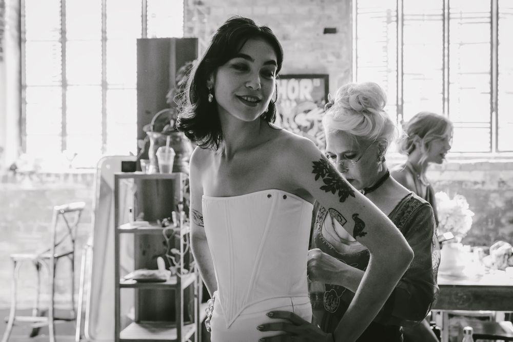 Black and white photo of a woman in a white corset being fitted by a tattooed stylist in a rustic studio.