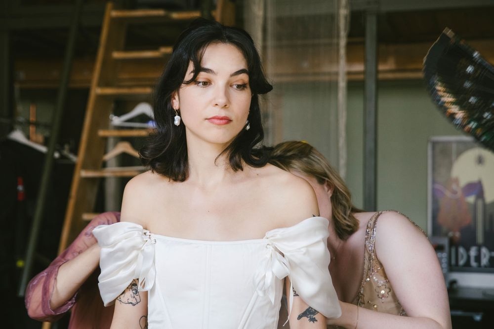 Woman in white off-shoulder bridal gown with tattoos and pearl earrings being fitted in a boutique.