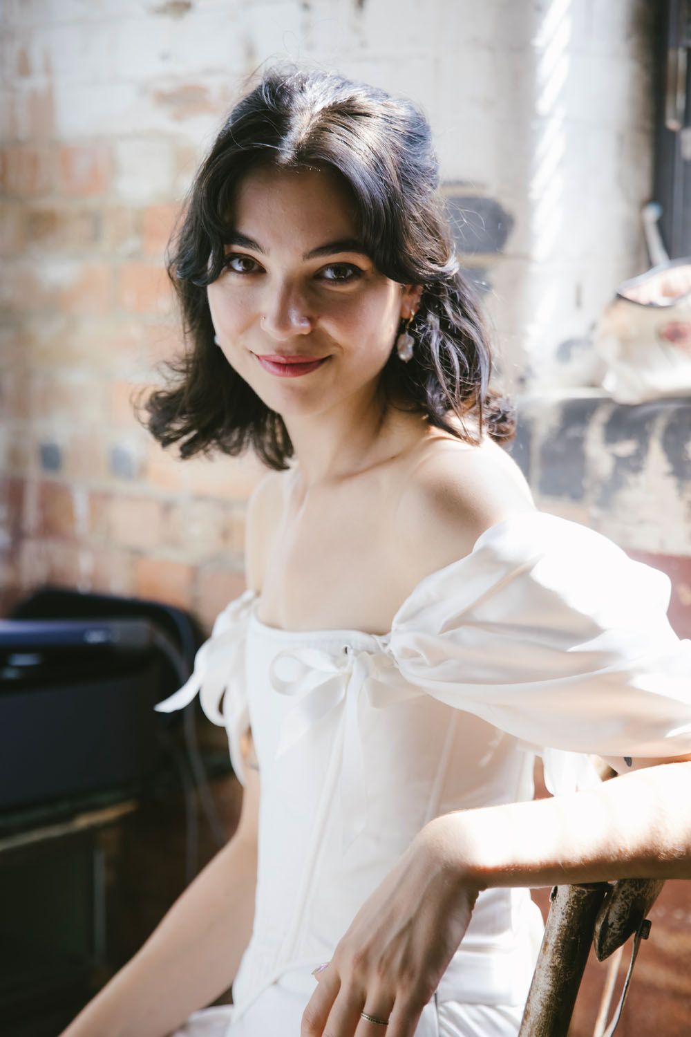 Young woman in white off-shoulder dress with bow detail poses gracefully in a rustic brick-walled studio.