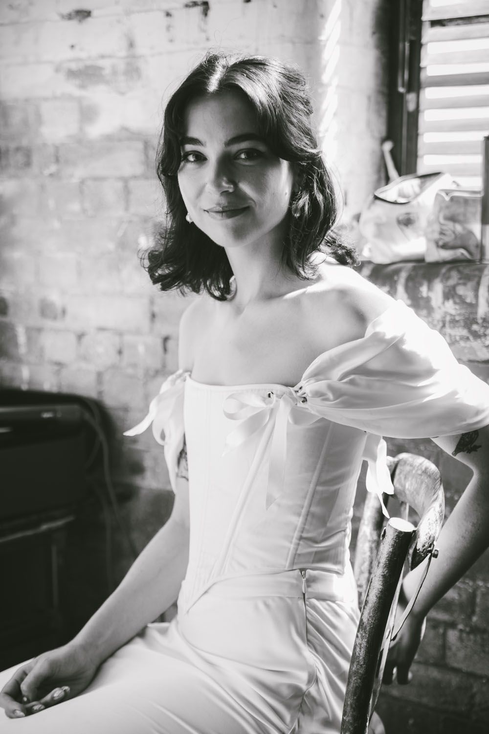 Black and white portrait of a young woman in a white off-shoulder corset dress seated in a vintage chair.