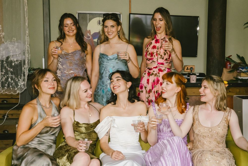 Bride in white dress celebrates with bridesmaids in colorful gowns, laughing and toasting with champagne glasses indoors.