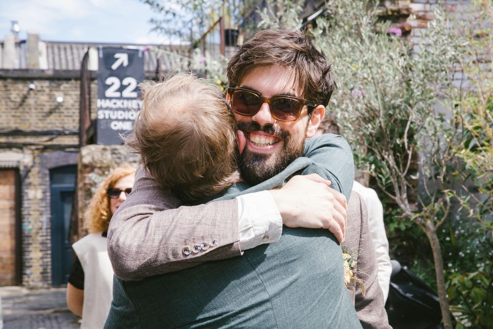 Two men sharing a warm hug outdoors near Hackney Studios on a sunny day, one wearing sunglasses and smiling.