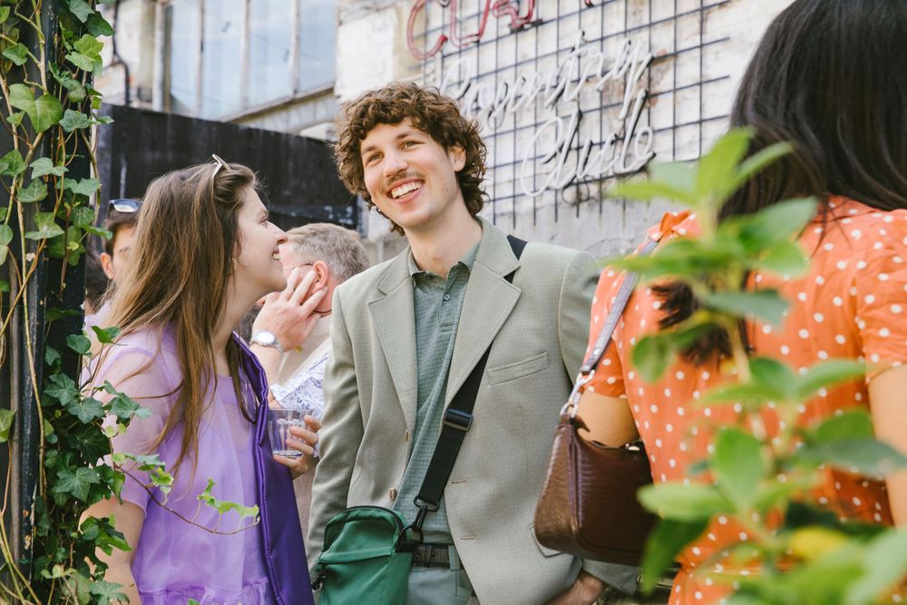 Smiling man in sage blazer chats with woman in purple dress at outdoor wedding reception.