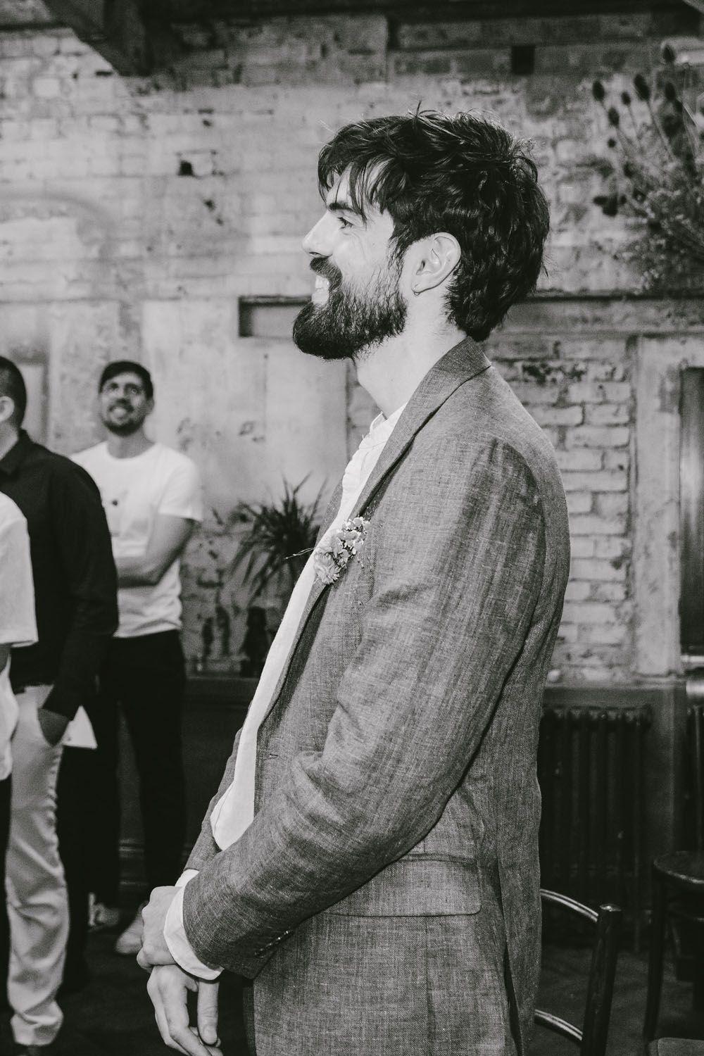 Bearded groom in gray linen suit smiles during wedding ceremony in rustic brick venue.
