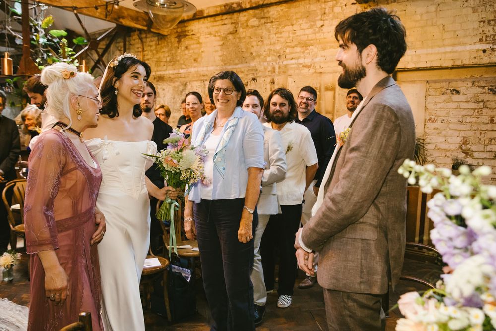 Bride in white gown greets guests with groom in brown suit at indoor wedding ceremony.
