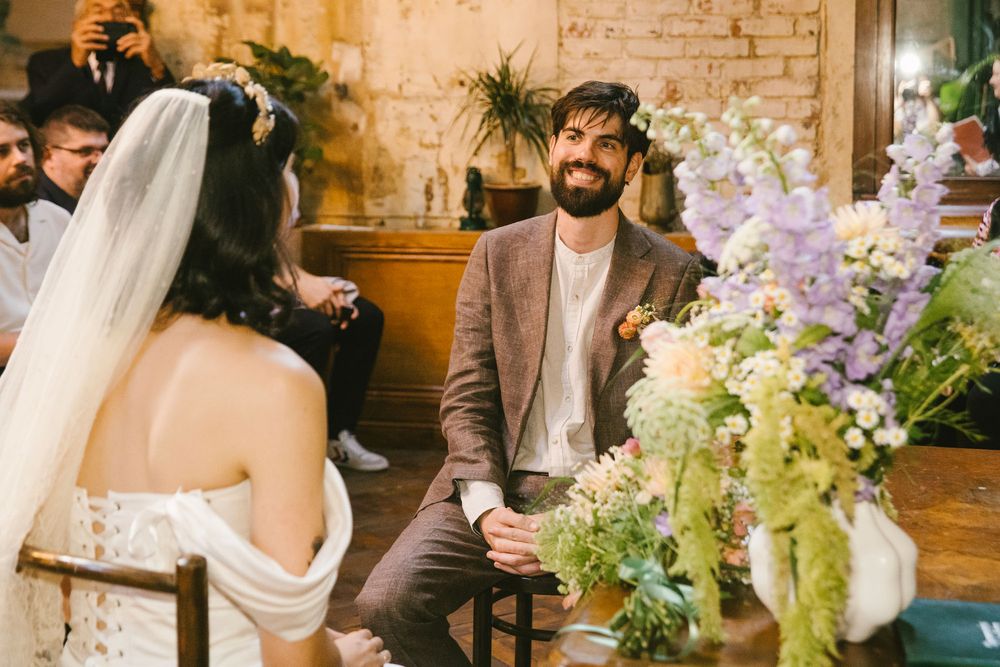 Groom in brown suit smiling at bride in white off-shoulder gown during indoor wedding ceremony with floral arrangements.