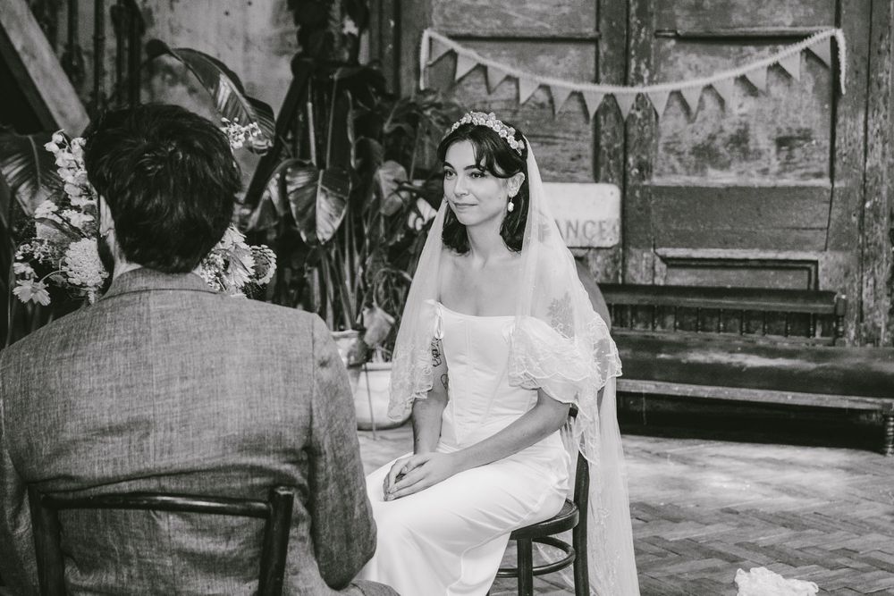 Bride in white dress and veil sitting during wedding ceremony in rustic venue, black and white photo.