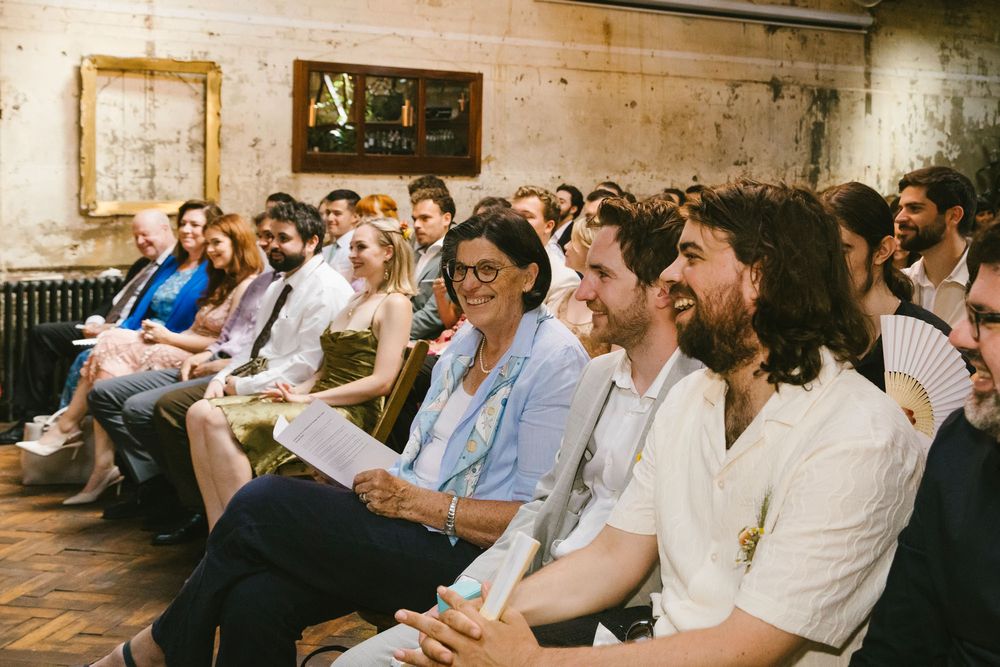 Wedding guests laughing and smiling during ceremony in rustic venue, candid color photograph.