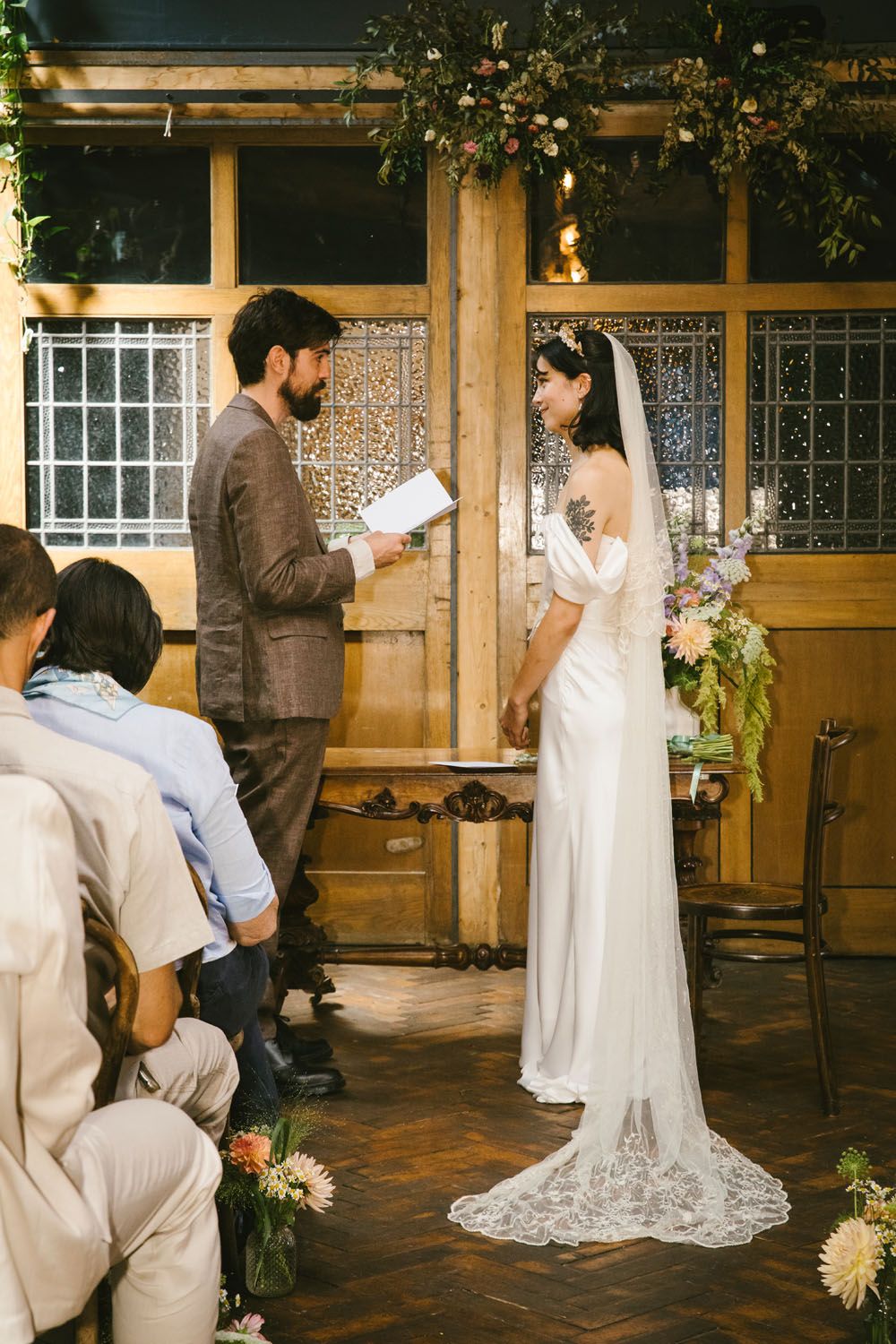 Bride and groom exchange vows at indoor wedding ceremony, bride in white lace gown with veil, groom in brown suit.