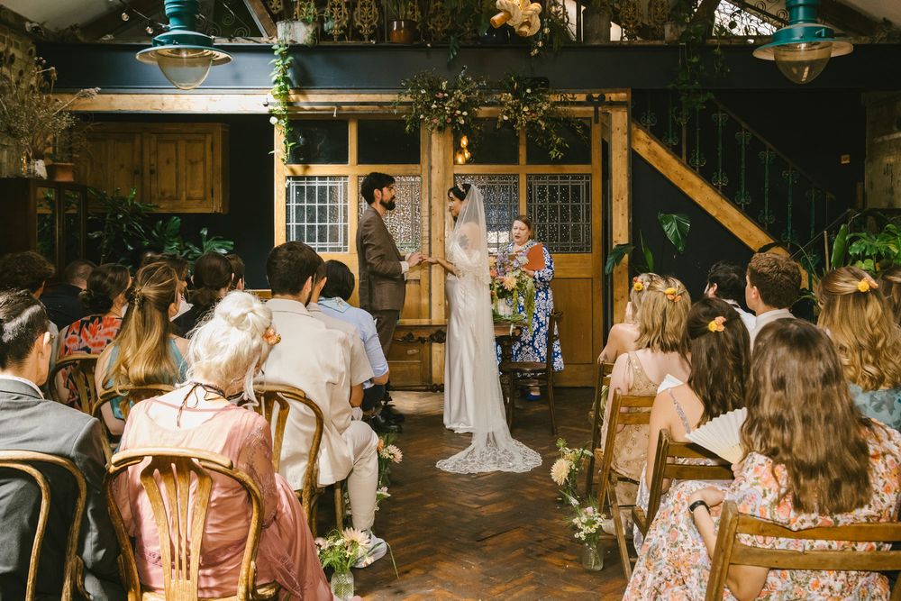 Indoor wedding ceremony with bride and groom at altar, guests seated in rustic venue with wooden beams and lush greenery decor.