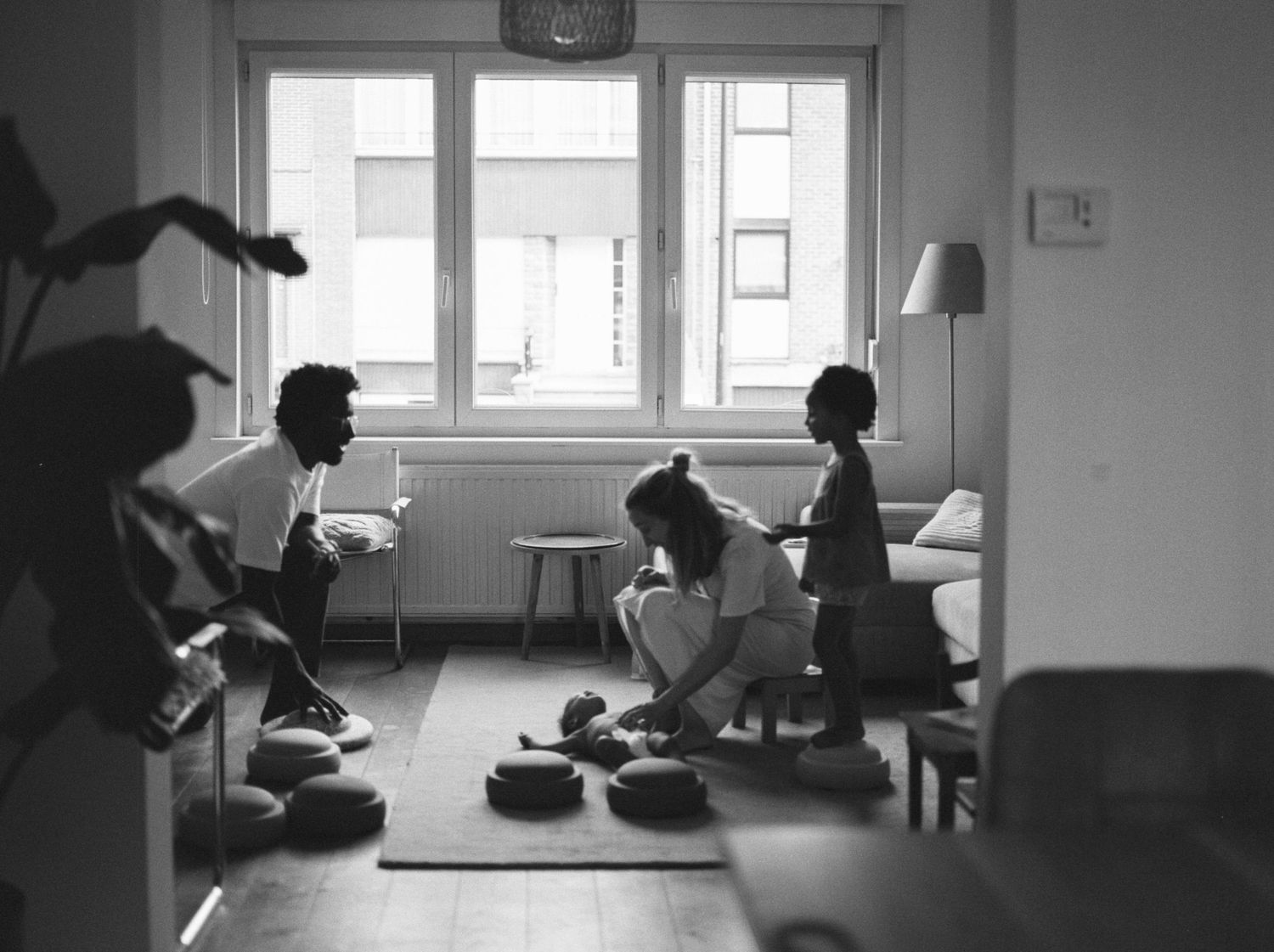 Black and white photo of three people relaxing in a bright living room, with cushions on the floor near a window.