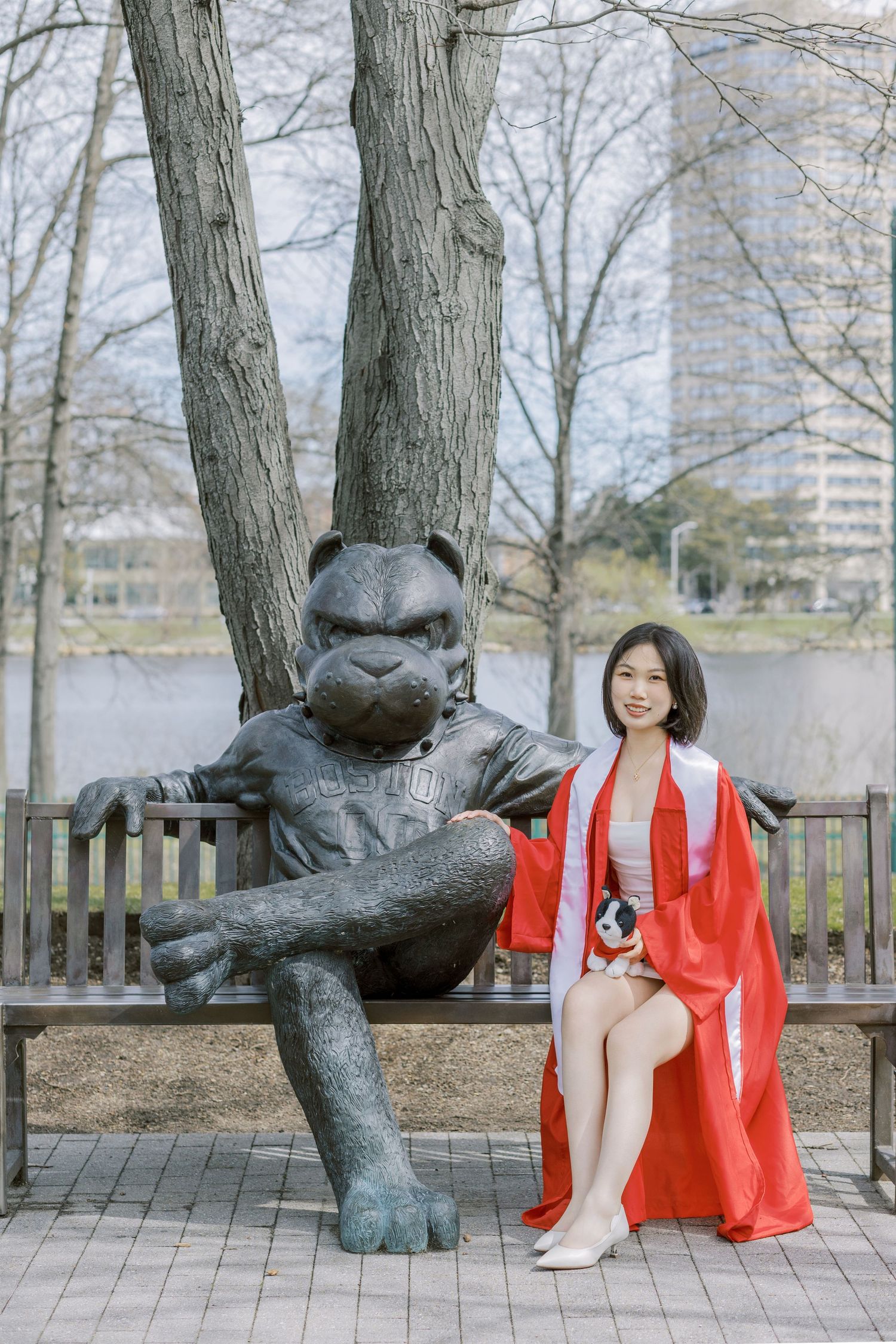 Boston University Graduate in red robe poses with bronze mascot statue on bench near river during spring graduation celebration.