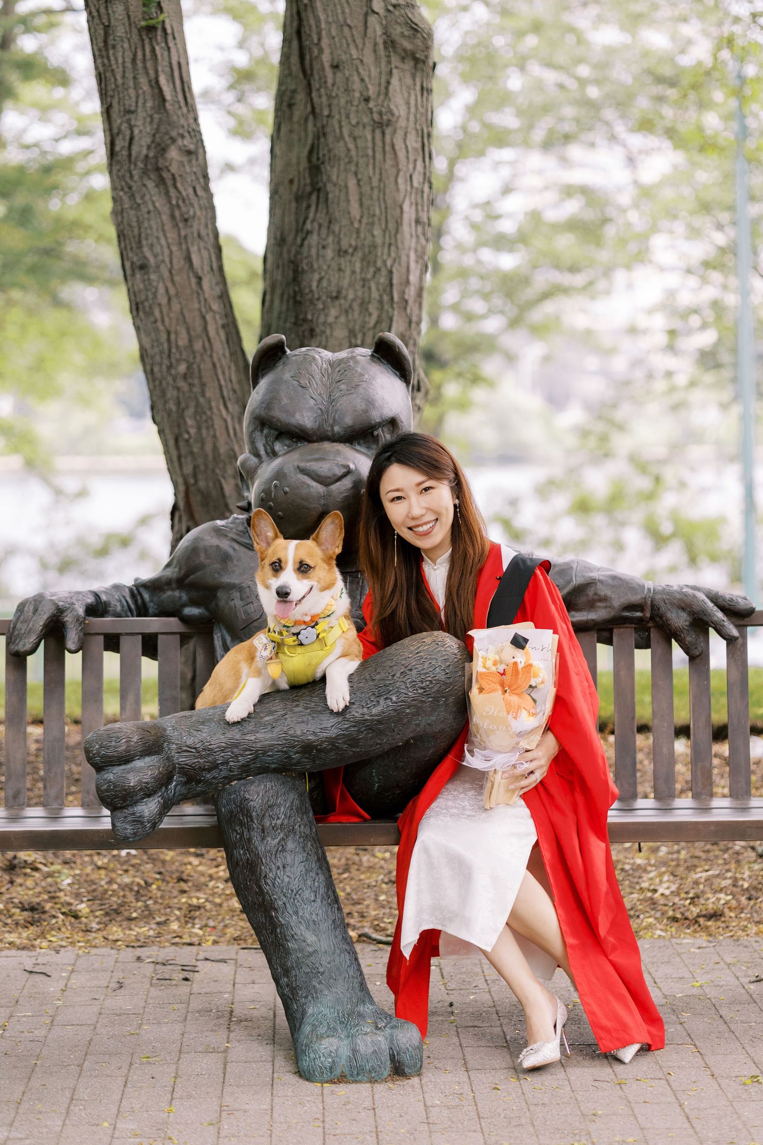 Smiling Boston University graduate in red robe holds bouquet and corgi beside bronze mascot statue on outdoor bench.