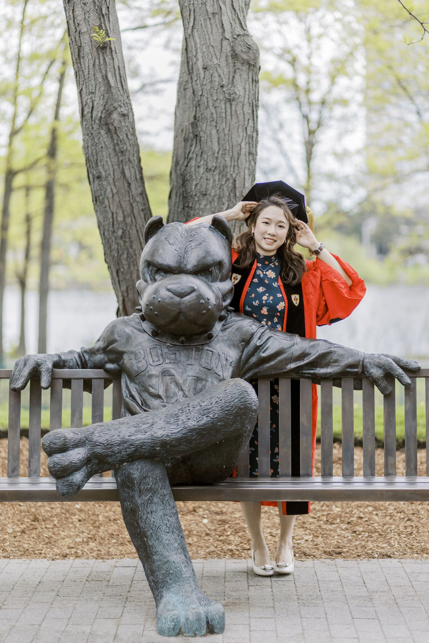 A smiling Boston University graduate in red gown and cap poses beside a bear mascot statue on a bench outdoors.