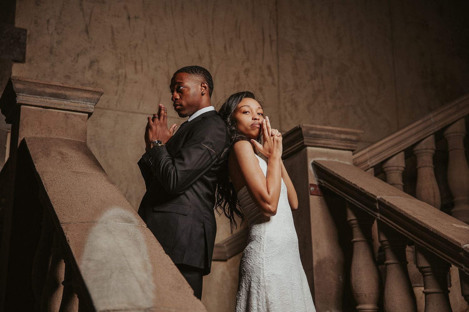 Elegant couple in wedding attire posing back-to-back on ornate staircase, groom in suit, bride in lace gown.