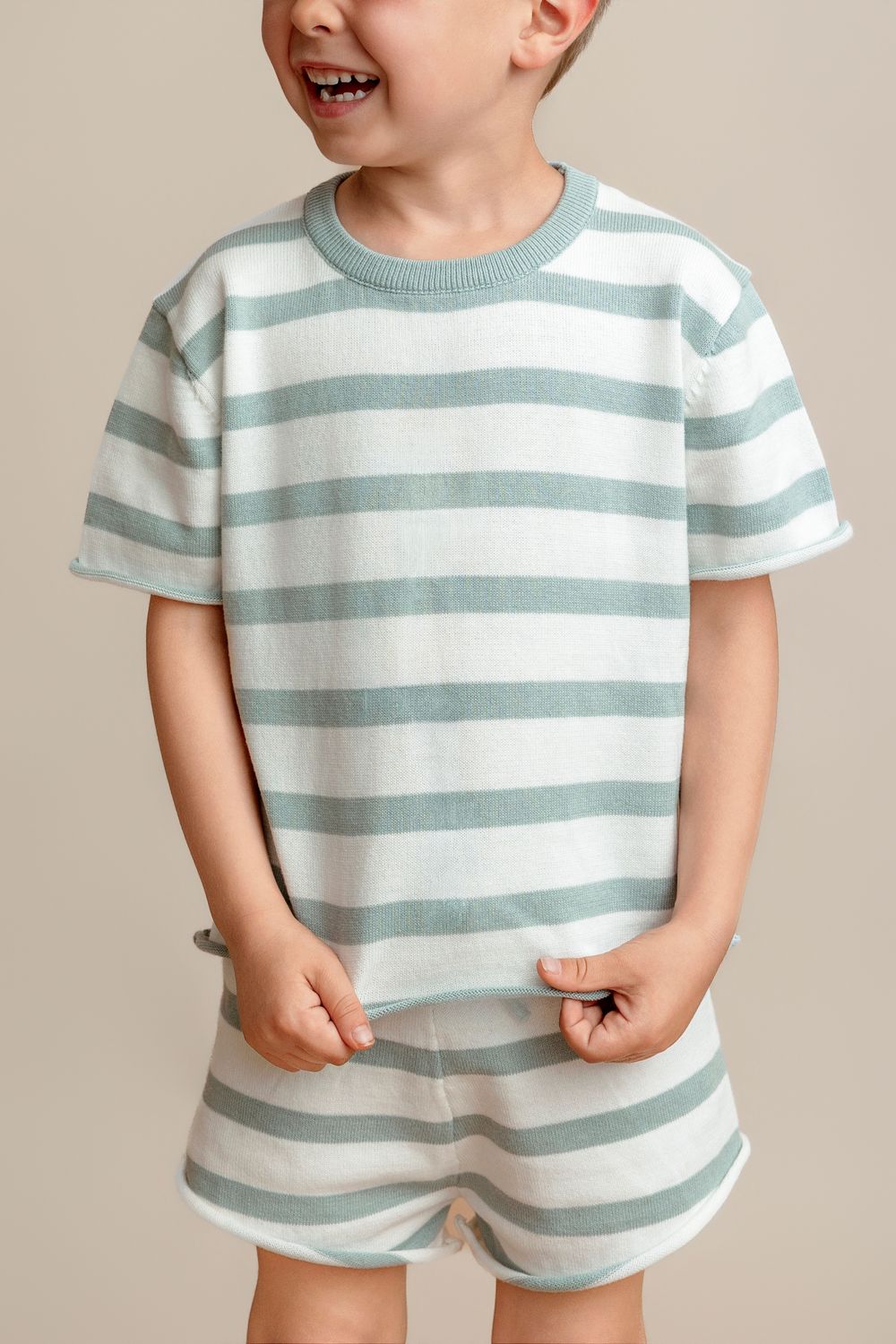 Smiling toddler boy wearing a sage green and white striped knit t-shirt and matching shorts set.