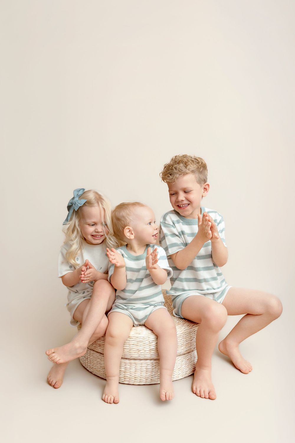 Three young siblings in matching striped outfits laugh and clap together on a woven ottoman against a neutral background.