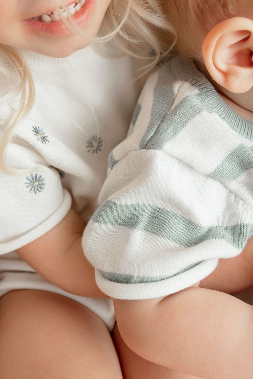 Two young siblings sitting together, older child in floral top hugging baby in sage green striped romper.