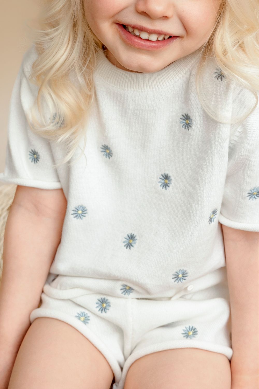 Smiling toddler girl wearing a white knit short set with blue daisy embroidery pattern.